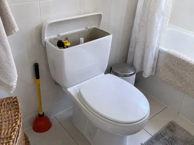 Baño de casa con el estanque del WC abierto y un desatascador apoyado en la pared
