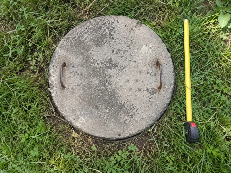 Tapa circular de hormigón de una fosa séptica en el pasto, junto a una huincha de medir